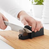 Person sharpens knife on black device on wooden board. Knife sharpener in kitchen.