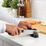 Person sharpens knife on device in kitchen, with ingredients nearby.