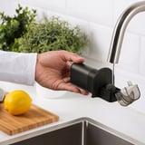 Person holding a black knife sharpener under a running faucet. The sharpener is filled with water.