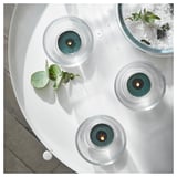 Three blue AVMÅLA scented candles on a white round table, each lit, with green leaves beside them.