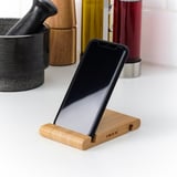 A black smartphone rests on a wooden stand with ikea engraved, positioned upright on a white surface, surrounded by decorative items.