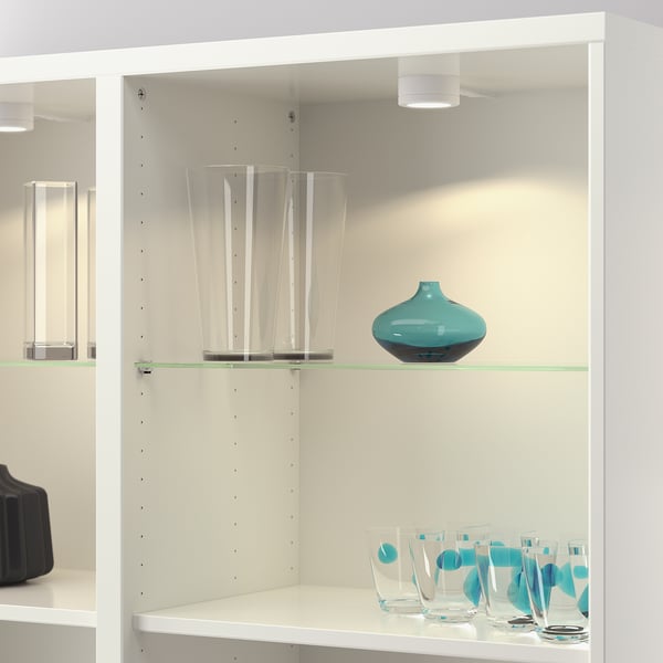 White shelf unit with glass shelves, holding decorative vases and patterned glasses. Glasses are placed on lower shelf, and vases on upper shelf.