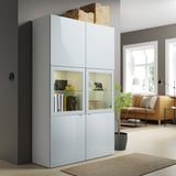 Light blue BESTÅ cabinet with glass doors, decorative items, near window & couch.