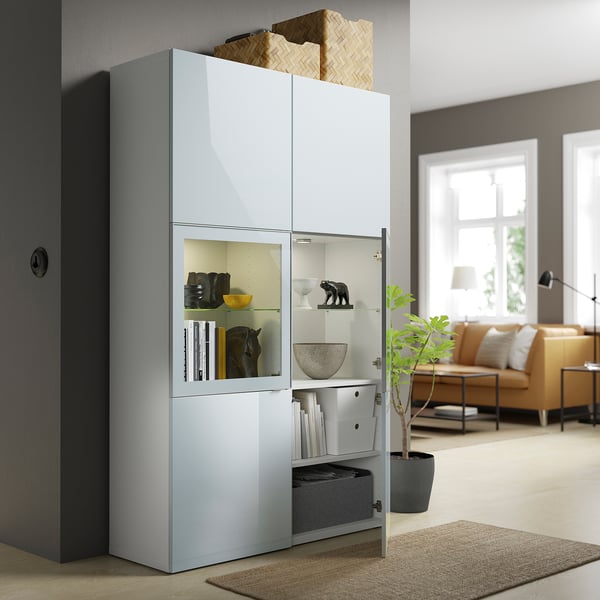 A light blue BESTÅ storage unit with glass doors showcasing books and decorative items, housed in a living area with a beige sofa and large window.