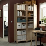 A wooden BESTÅ bookcase stores various storage boxes, books, and items. The shelves are adjustable, showcasing organizational versatility.