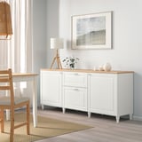 White BESTÅ sideboard with adjustable shelves, drawers, light wooden top, seascape art, wooden chair, curtained window.