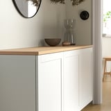 Minimalist white BESTÅ sideboard with oak top, featuring decorative bowl and vase. Mirror and stool nearby for context.
