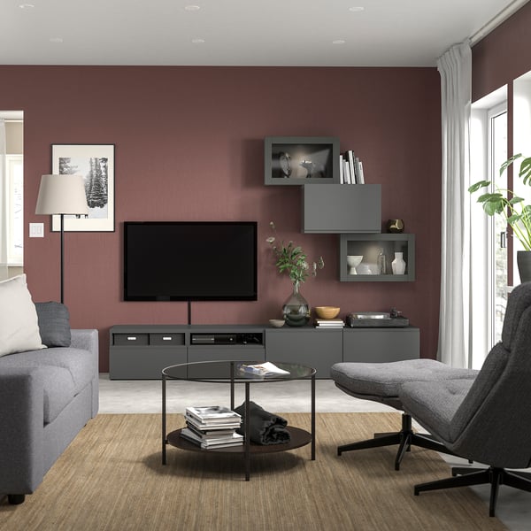 Modern living room with maroon walls, grey BESTÅ tv unit, grey couch, glass coffee table, black & grey lounge chair, white curtains, wooden floor.