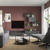 Modern living room with maroon walls, grey BESTÅ tv unit, grey couch, glass coffee table, black & grey lounge chair, white curtains, wooden floor.