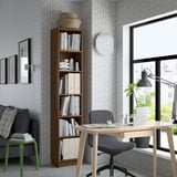 A well-organized home office with a wooden bookcase, office chair at a desk by a window with a laptop and plant, and a cozy sofa with pillows.