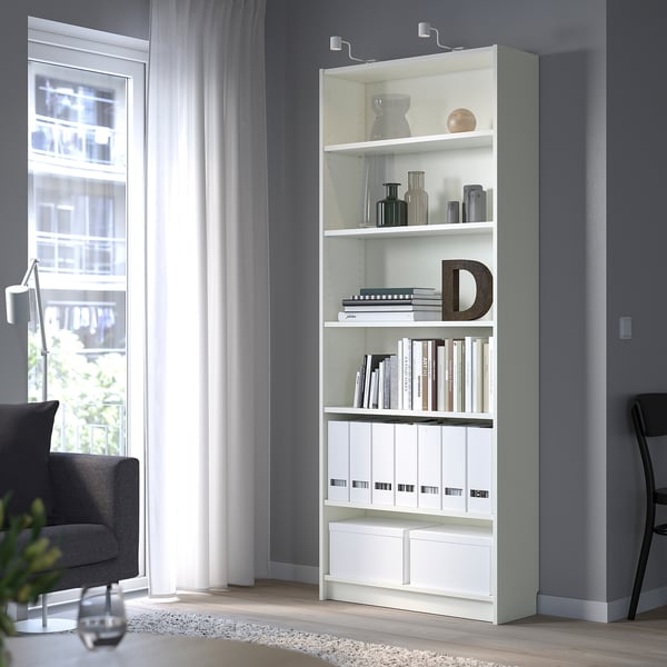 White BILLY bookshelf with books, vases, & letter d. Next to window & grey chair, adjustable shelves, particleboard & melamine, bright room.