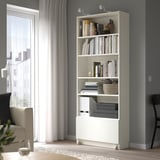 White BILLY bookcase by window, filled with books, vase, basket, and drawer. Towering over armchair and side table, with curtains framing the window.