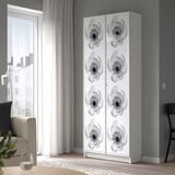Modern living room with tall white BILLY bookcase, adorned with black floral designs, standing near large window with sheer curtains.