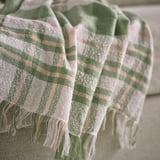 Textured, striped throw blanket with fringe in green and cream resting on a sofa. Soft, lightweight weave visible.