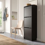 Black shoe cabinet with 3 compartments next to a beige chair and narrow wall unit with jackets.