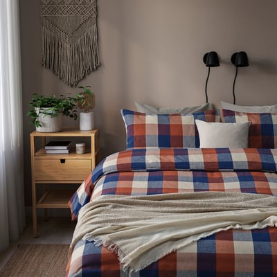 Bed with colorful checkered BLÅSENAP duvet, soft cotton, wall hangings, wooden nightstand holding plants and books.