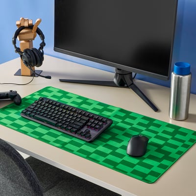 Gaming setup with large green checkered BLÅSKATA mouse pad. Keyboard, mouse, monitor, headphones, and water bottle on a grey desk.