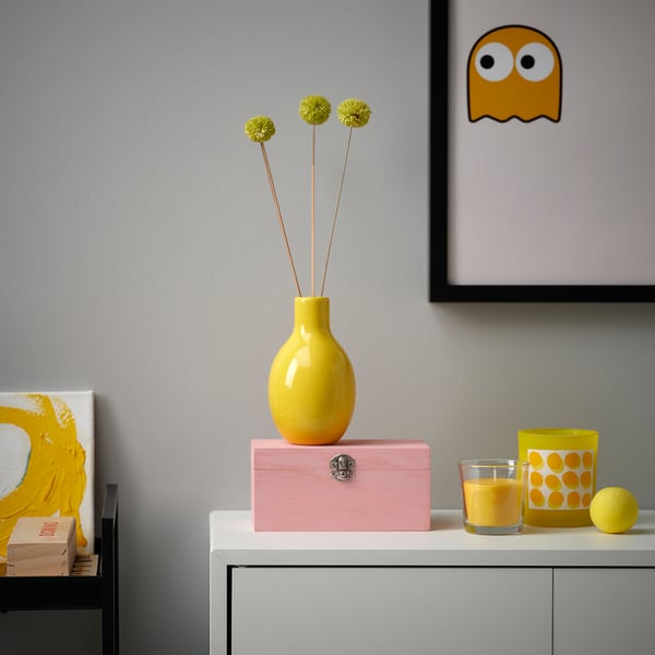 Bright yellow BLODBJÖRK vase with round shape holds three flowers. Placed on pink box near glassware and artwork, highlights cheerful decor.