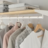 A closet with wooden and white hangers holding clothes like sweaters and a jacket. Folded blankets are stored on a shelf above.