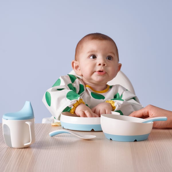 Baby wearing green polka dot bib sits at table with BÖRJA spoon set: one spoon for self-feeding, another for jar use.