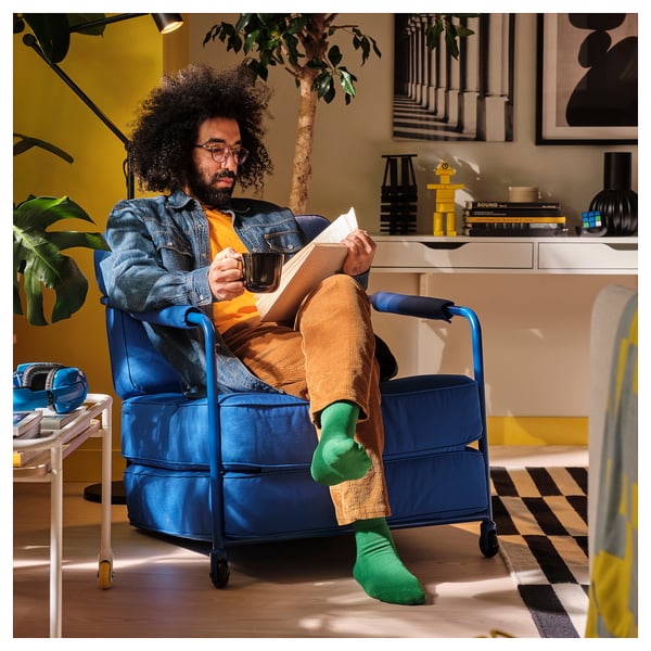 Man sits in a blue BRÄNNBOLL chair, holding a mug, reading. Chair a movable seat making it versatile for reading or lounging.