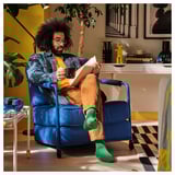 Man sits in a blue BRÄNNBOLL chair, holding a mug, reading. Chair a movable seat making it versatile for reading or lounging.