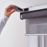 A hand holding a small black device connects it to a motorized roller blind, demonstrating its use to transfer back-up power.