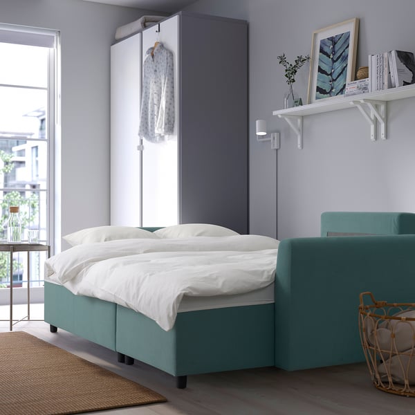 Turquoise sofa-bed in compact bedroom with white bed, green armchair, and small shelf beside window.