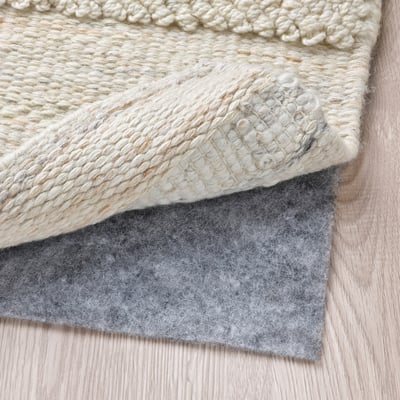 A close-up of a beige woven rug laid on a light wooden floor, with gray felt underlay visible, showcasing its texture and functional placement.