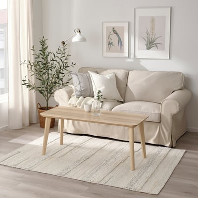 Minimalist living room: light beige BRÖNDEN rug, natural wood coffee table, cream sofa, white walls, large plant, nature prints.
