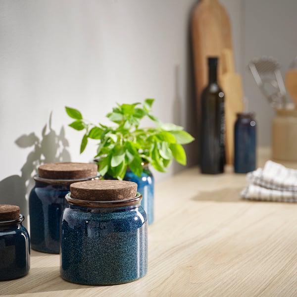BRUGDHAJ blue jars with cork lids on wooden table. Near green plant, bottles, and utensils. Medium-sized, rustic, glazed stoneware with cork tops.