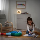 Little girl sits on rug, reading a book by BRUMMIG led lamp. Soft light illuminates room, showcasing a cozy space with plush toys.