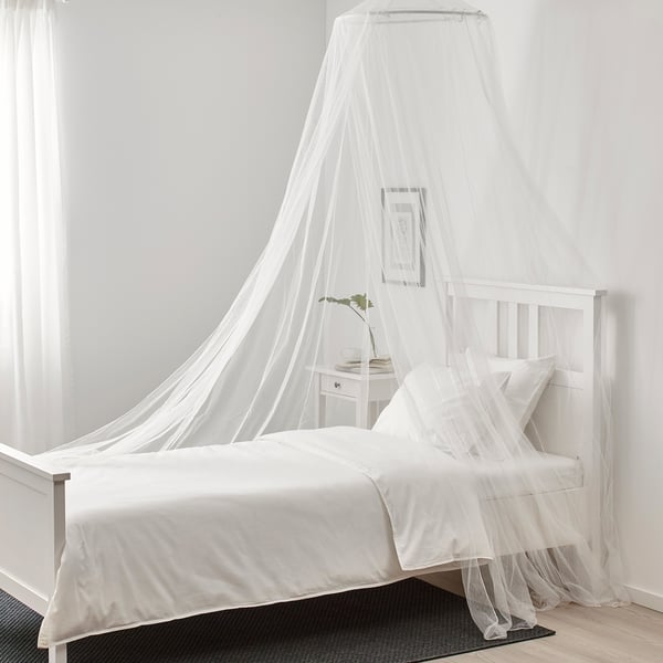 White canopy bed with net, plastic frame, easy-clean polyester cover. Beside nightstand with plant, picture, black rug.