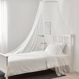 White canopy bed with net, plastic frame, easy-clean polyester cover. Beside nightstand with plant, picture, black rug.