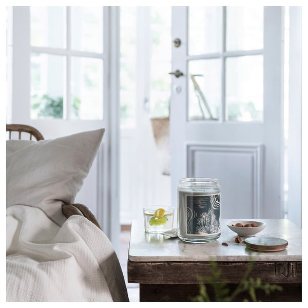 Scented candle in glass, white label, beside chair by window with blanket.