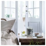 Scented candle in glass, white label, beside chair by window with blanket.