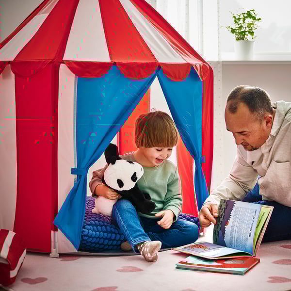 A child sits inside a colorful play tent with a man reading a book, illustrating a cozy hiding spot where children can play or listen to stories.