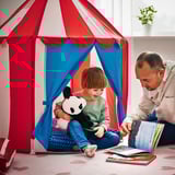 A child sits inside a colorful play tent with a man reading a book, illustrating a cozy hiding spot where children can play or listen to stories.