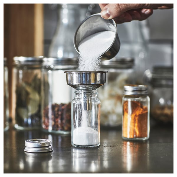 A hand pours white substance into clear glass jar with metal funnel top from metallic container. Surrounded by similar jars and bottles in a kitchen.