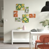 Colorful nature-inspired art in orange and green on wall above white table with pink vase and fruit.
