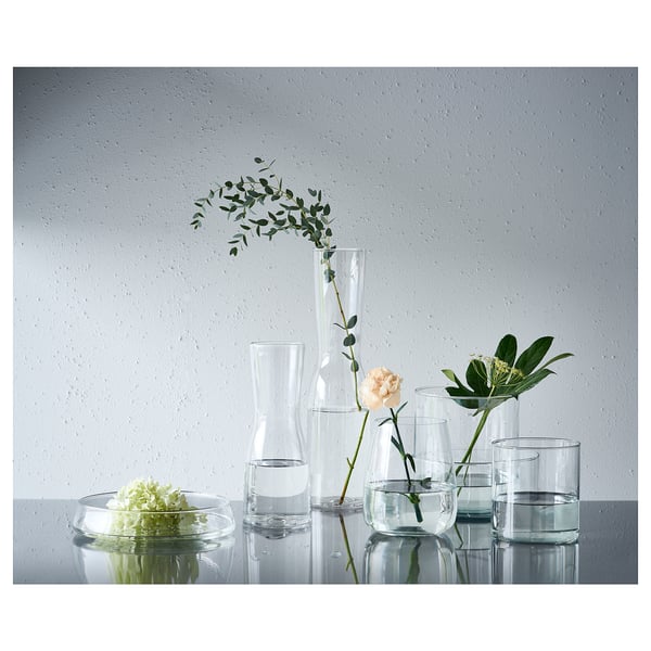 Six clear glass vases of varying sizes display flowers on a white surface against a gray wall.