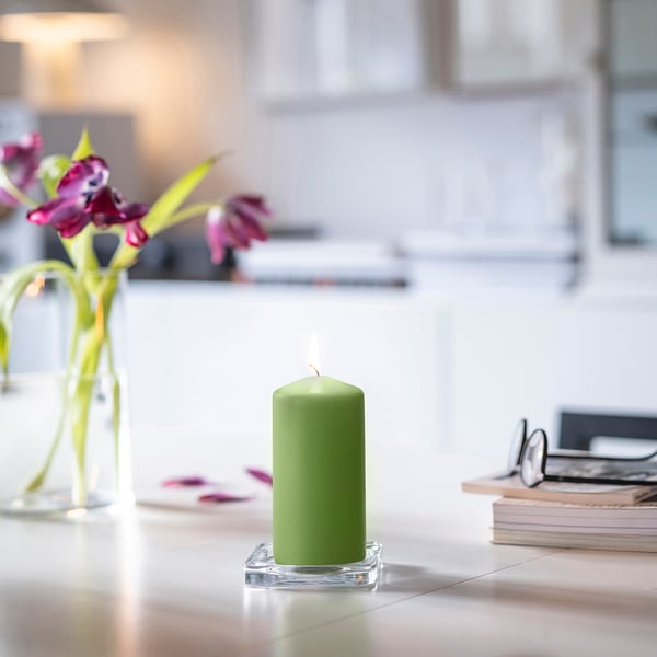 Small lit green DAGLIGEN candle on clear holder beside pink flowers in vase, glasses on books.