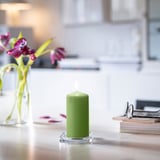 Small lit green DAGLIGEN candle on clear holder beside pink flowers in vase, glasses on books.