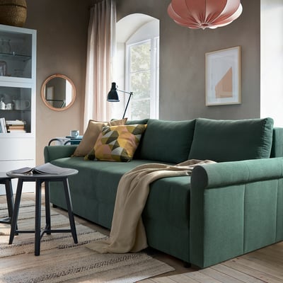 A cozy living area with a DÅNHULT green sofa bed, decorated with patterned pillows, situated near a window. A small side table holds a mug and book.