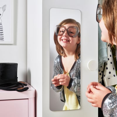 A young girl styles herself in a mirror attached to a wardrobe door.