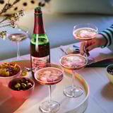 Three cocktail glasses with pink drinks, a bottle labeled DRYCK BUBBEL ÄPPLE & LINGON, and bowls of snacks on a table.