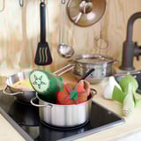 Kids play kitchen with metal pots, green, red, and beige mittens. Wooden backdrop, hanging utensils, black faucet.