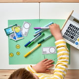 Child draws on play money with markers. Calculator and striped shirt visible.