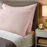 Two soft pink DVALA cotton pillowcases on a bed. Theyre rectangular with smooth edges, larger than the bedside clock and lamp.