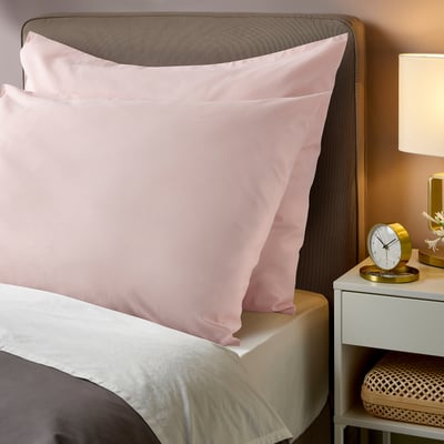Two soft pink DVALA cotton pillowcases on a bed. Theyre rectangular with smooth edges, larger than the bedside clock and lamp.